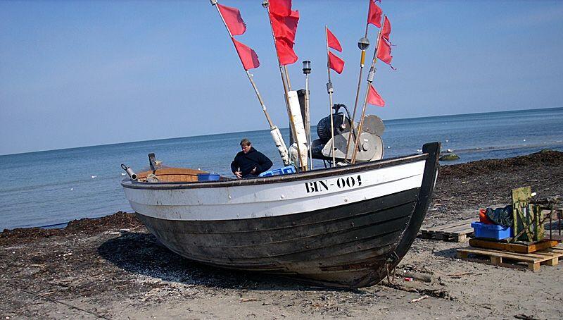 Kutter mit roten Fangflaggen: Ein traditionelles Fischerboot mit der Kennzeichnung BIN-004 steht auf einem Strandabschnitt. Am Boot sind zahlreiche rote Fangflaggen montiert, im Hintergrund ist die ruhige Ostsee zu sehen.