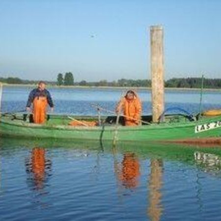 Zwei Fischer in orangefarbener Kleidung stehen in einem kleinen grünen Holzboot auf einem ruhigen See.