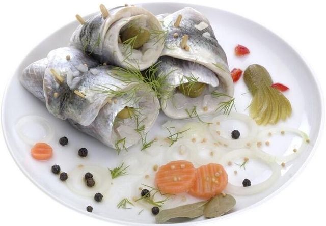 Drei Rollmöpse auf einem weißen Teller, garniert mit Zwiebeln, Gurke, Möhrenscheiben, Pfefferkörnern, Senfkörnern und Dill.